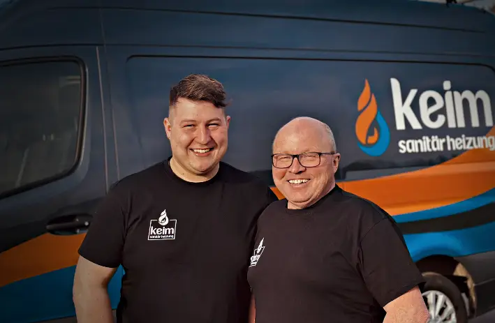 Das Foto zeigt Alfred und Christoph, Vater und Sohn, die stolz ihr Familienunternehmen „Keim Sanitär Heizung“ repräsentieren. Alfred, der ältere von beiden, steht mit einer Haltung der Erfahrung und Zuversicht links im Bild. Er trägt Arbeitskleidung, die sauber und professionell aussieht, symbolisch für jahrzehntelange Hingabe und Meisterschaft in seinem Handwerk. Sein Gesichtsausdruck strahlt Stolz und ein tiefes Verständnis für sein Handwerk aus. Christoph, rechts neben seinem Vater, trägt ähnliche Arbeitskleidung, die die Tradition und das Fortführen des Familienbetriebs unterstreicht. Sein jugendlicher Enthusiasmus und seine Entschlossenheit sind in seinem motivierten Blick erkennbar. Beide stehen vor dem Hintergrund ihres Betriebs – vielleicht einem Lager voller Werkzeuge und Materialien oder einem sauber installierten Heizungssystem – was ihre Fachkenntnisse und die Qualität ihrer Arbeit hervorhebt. Die Verbindung zwischen Vater und Sohn ist durch ihre ähnliche Körperhaltung und die Art, wie sie zueinander stehen – nah, doch selbstständig – symbolisiert, was Respekt, Zusammenarbeit und die Übergabe von Werten und Wissen von einer Generation zur nächsten ausdrückt. Das Licht im Bild fokussiert auf ihre Gesichter und die Details ihrer Arbeitskleidung, was ihre Rollen als Handwerker und Hüter familiärer Traditionen betont.